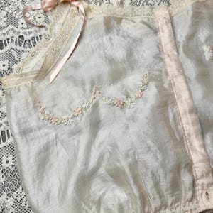 May include: A delicate white silk camisole with a pink satin ribbon and lace trim. The camisole features a floral embroidery design in pink and white.