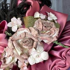 May include: A pink satin hat with a black feather and a cluster of pink and white fabric flowers.