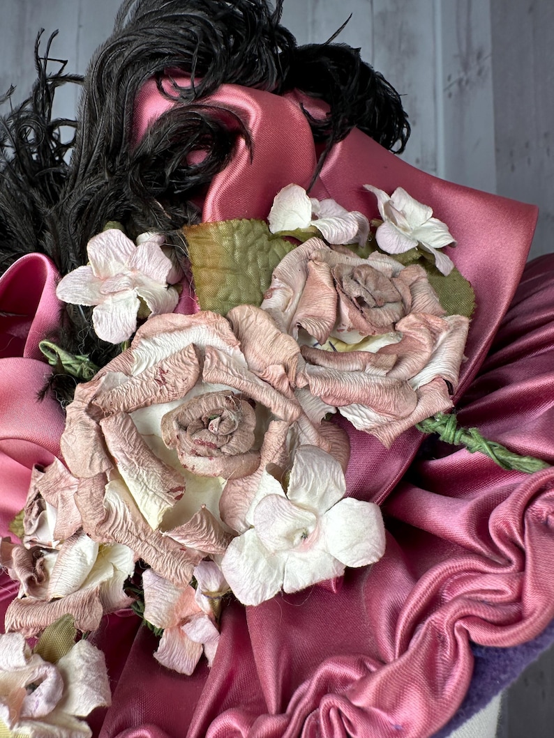 May include: A pink satin hat with a black feather and a cluster of pink and white fabric flowers.