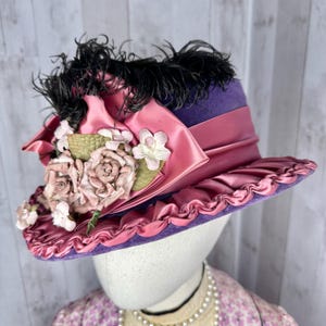 May include: A purple velvet hat with a pink satin ribbon and bow, adorned with pink fabric flowers and black feathers.