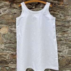 May include: A white sleeveless chemise featuring a square neckline with delicate lace trim along the edges and shoulder straps. The garment is made of a smooth, solid white fabric and hangs straight down from a wooden hanger.
