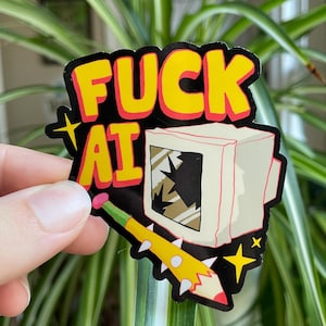 Könnte beinhalten: Ein Aufkleber mit den Worten "FUCK AT" in gelben Buchstaben mit roter Umrandung, vor schwarzem Hintergrund. Der Aufkleber zeigt ein Cartoon-Bild eines kaputten Computermonitors und einen gelben Bleistift mit weißen Spitzen.