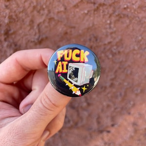 Op de afbeelding: Een zwarte knoop met de tekst "FUCK AI" in geel en rood. De knoop heeft een retro computer graphic en een cartoon raket. De knoop is rond en heeft een glanzende afwerking.