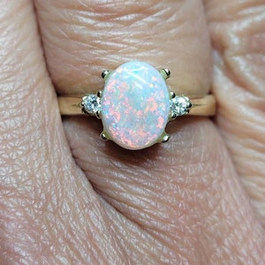 May include: A gold ring with an oval-shaped opal gemstone and two smaller diamond accents. The opal has a white base with flashes of pink and blue.