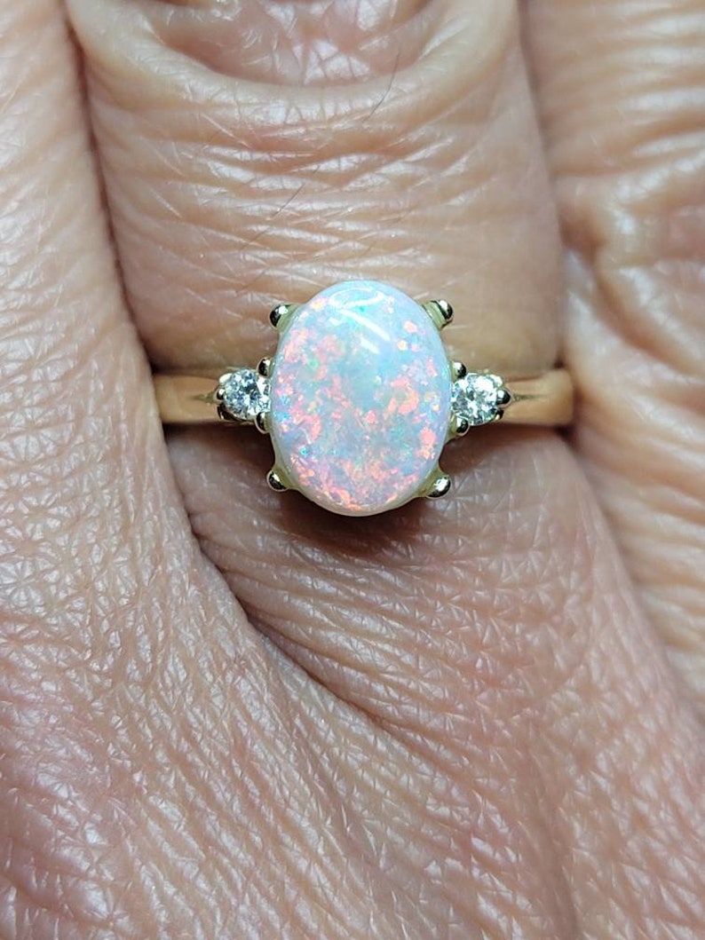 May include: A gold ring with an oval-shaped opal gemstone and two smaller diamond accents. The opal has a white base with flashes of pink and blue.