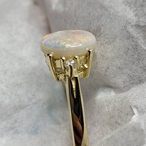 May include: A gold ring with a white opal gemstone and a small diamond accent. The ring has a delicate, ornate design.