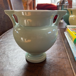 May include: A light blue ceramic vase with two handles. The vase has a rounded body and a pedestal base. The handles are rectangular and extend outward from the rim. The vase is sitting on a brown surface.