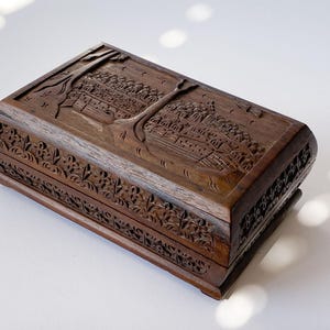 May include: A rectangular wooden box with intricate carvings. The box features a detailed landscape scene on the lid, including trees and buildings. The sides of the box are adorned with floral patterns. The wood has a rich, dark brown color.