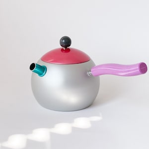 Puede incluir: Una tetera moderna de color plateado con tapa rosa y asa morada. El pico es turquesa y la tapa tiene un pequeño pomo gris oscuro. La tetera es redonda y está sobre una superficie blanca.