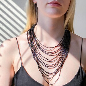 May include: A layered necklace made of iridescent black beads. The necklace is worn by a person with long blonde hair and is styled in a V-shape.