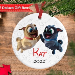 May include: A white round Christmas ornament featuring two cartoon pugs from the movie "The Secret Life of Pets". The pugs are in mid-air, one is brown and the other is gray. The ornament is personalized with the name "KAT" and the year "2022" in red.