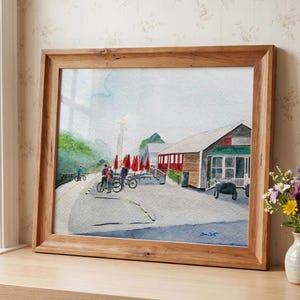 May include: A framed watercolour painting depicting a roadside scene with a building, red umbrellas, and people with bicycles on a paved path. The artwork features soft blue, green, and red tones, signed in the bottom right corner, and housed in a wooden frame.