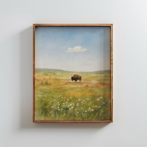May include: A framed landscape painting featuring a brown bison standing in a vast, grassy field filled with small white wildflowers under a pale blue sky with a single white cloud. The artwork is enclosed in a simple, medium-toned wooden frame.