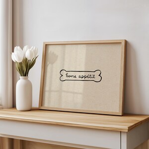 May include: A framed print with a dog bone design and the words "bone app&eacute;tit" in black script. The print is in a light wood frame and rests on a wooden surface. A white vase with white tulips is to the left.