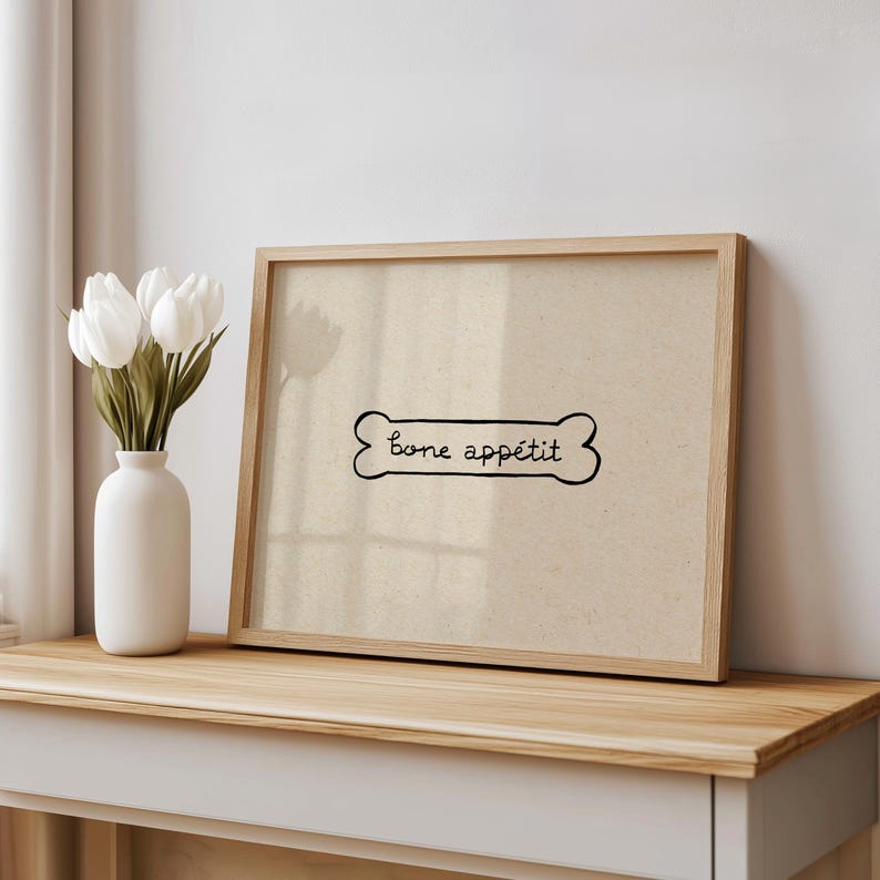 May include: A framed print with a dog bone design and the words "bone app&eacute;tit" in black script. The print is in a light wood frame and rests on a wooden surface. A white vase with white tulips is to the left.