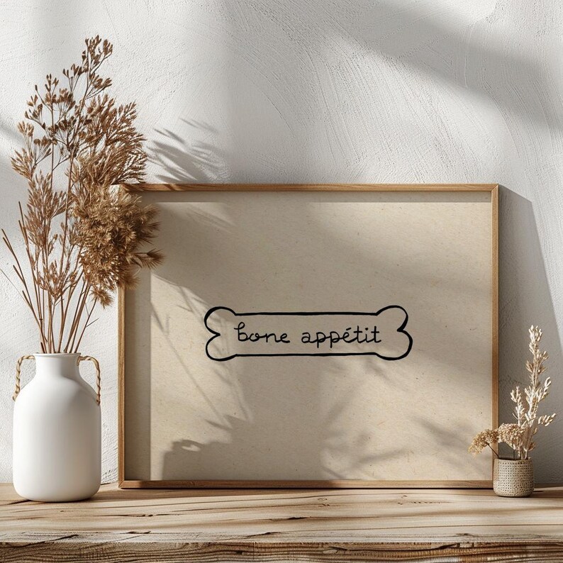 May include: A framed print with the words "Bone app&eacute;tit" in a playful font, designed as a dog bone. The print has a neutral color palette and is displayed on a wooden surface. Decorative elements include a white vase with dried plants.