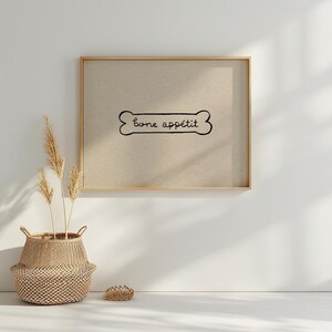 May include: A framed print with the words "bone app&eacute;tit" in a bone-shaped design. The print is in a gold frame and hangs on a white wall. A woven basket with dried plants sits below the print.