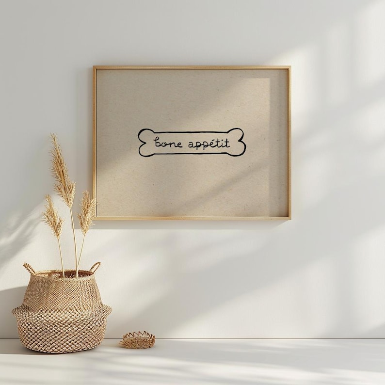 May include: A framed print with the words "bone app&eacute;tit" in a bone-shaped design. The print is in a gold frame and hangs on a white wall. A woven basket with dried plants sits below the print.