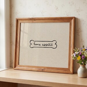 May include: A framed print with a light brown background and a wooden frame. The print features a black bone shape with the words "bone app&eacute;tit" written inside. A small vase of flowers sits to the right.