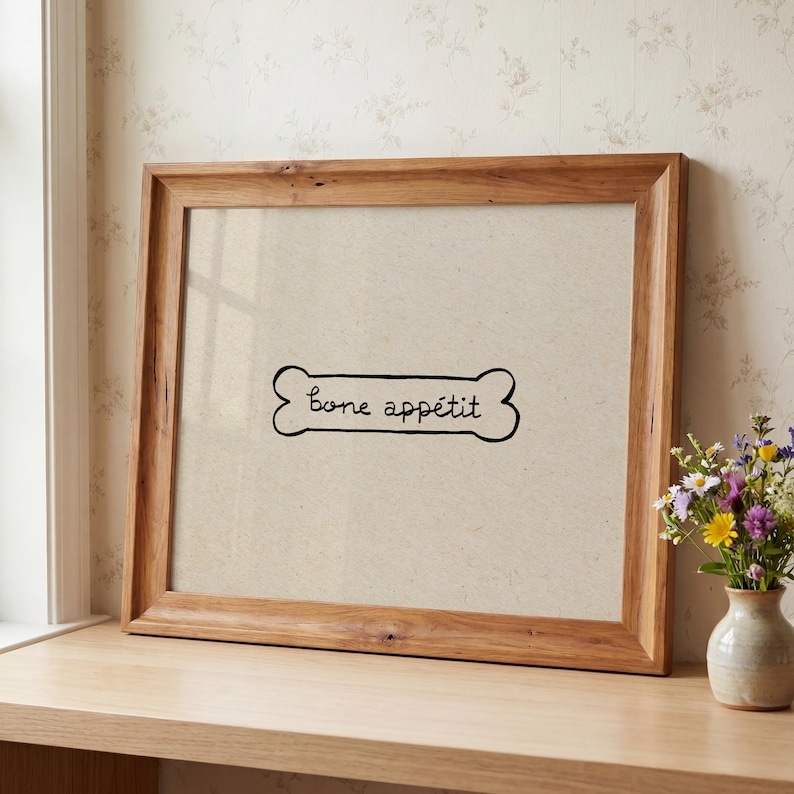 May include: A framed print with a light brown background and a wooden frame. The print features a black bone shape with the words "bone app&eacute;tit" written inside. A small vase of flowers sits to the right.