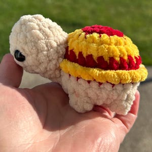 May include: A small crocheted turtle toy with a white body and a red and yellow shell.