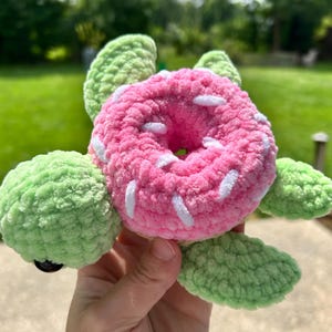 May include: A handmade crochet turtle with a pink donut shell and green flippers. The donut has white sprinkles. The turtle is a unique, whimsical creation.