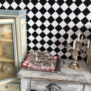 Pode incluir: Um conjunto de chá prateado em miniatura numa bandeja decorativa com uma toalha de mesa xadrez vermelha e branca. O conjunto inclui uma chaleira, um creme, um açucareiro e duas chávenas de chá com pires. Um castiçal prateado com velas brancas.