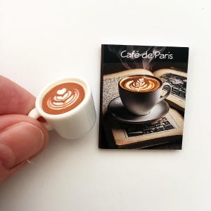 May include: A white ceramic coffee mug with a latte art design. The mug is sitting on a hand. The background is a close-up of a book with the text "Café de Paris" on the cover.