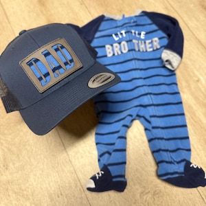 May include: A navy blue trucker hat with a leather patch that reads "DAD" in blue stripes. Next to the hat is a blue striped baby onesie with the words "LITTLE BROTHER". The onesie has attached feet.