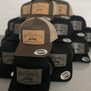 May include: A collection of trucker hats in various colors, including black and brown. Each hat features a rectangular patch with the text "Sheriff-Goslin Co. ROOFING SINCE 1906" and a graphic of a truck. The hats have a mesh back and a snapback closure.
