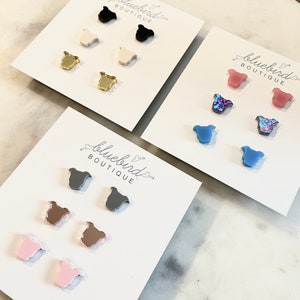 May include: Multiple sets of stud earrings in various colors, including black, gold, pink, and blue, are displayed on white cards. The earrings are shaped like a stylized bird. The cards are labeled "Bluebird Boutique."