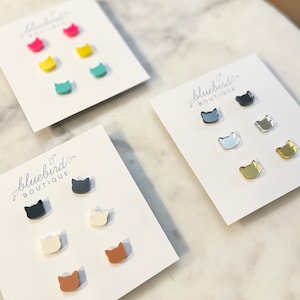 May include: Three sets of cat-shaped stud earrings on white cards. The first set is black, white, and brown. The second set is silver, light blue, and gold. The third set is pink, yellow, and turquoise.