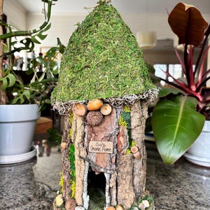 以下が含まれることがあります： 小枝と苔でできたミニチュアの妖精の家。看板には「Owl's Gnome Home」と書かれています。