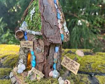 May include: A miniature fairy house crafted from bark and twigs, adorned with moss, seashells, and glass beads. The house features a sign that reads "Though she be but little, she is fierce!"