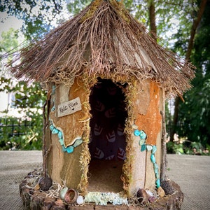 Puede incluir: Una pequeña casa de hadas rústica hecha de madera y ramitas con un techo de paja. La casa tiene una pequeña puerta con un letrero que dice "Kale Fries". La casa está decorada con musgo, piedras y otros elementos naturales.