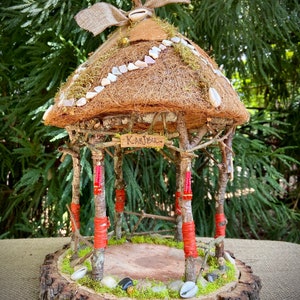 Puede incluir: Una casa de hadas artesanal con techo de paja, adornada con conchas marinas y musgo. La estructura de madera presenta postes envueltos en rojo y una base de tronco de árbol. El letrero dice "Karibu".