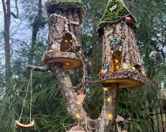 May include: A miniature fairy house with two levels, built from tree bark and moss, with a swing made from a halved coconut shell. The house is decorated with small stones, acorns, and fairy lights. The base is a wooden slice covered in moss.