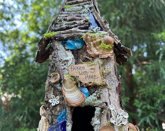 May include: A miniature fairy house crafted from twigs and bark, adorned with blue glass, acorns, and a sign that reads "Fairies Live Here".