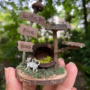 May include: A miniature fairy house made of natural materials, including wood, moss, and acorns. The house has a small sign that reads "Little Bit", and another sign that reads "Blue Hoss". A small silver cat figurine sits in front of the house.
