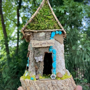 May include: A miniature fairy house made of wood and moss with a sign that reads "Love Lives Here". The house has a small door and is decorated with turquoise beads and a silver butterfly.