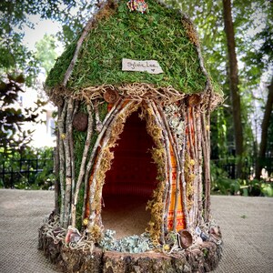 Puede incluir: Una pequeña casa de hadas cubierta de musgo, hecha de ramitas y ramas. La casa tiene una pequeña puerta y un letrero que dice "Sylvia Lee".