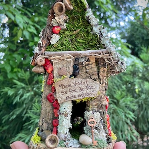 Puede incluir: Una casa de hadas en miniatura hecha de corteza y musgo con un letrero que dice "Bem Vindas Se for de Paz Pode Entrar". La casa tiene una pequeña puerta y una cerradura con una llave. La casa está sentada sobre una base de madera.