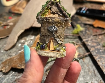 May include: A miniature fairy house made of bark and moss with a small wooden door and a metal roof. The house is being held in a hand.