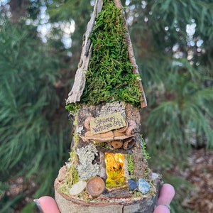 May include: A miniature fairy house crafted from wood and moss, featuring a sign that reads "Grumpy Witches Only". The house has a small window with a warm glow emanating from within.
