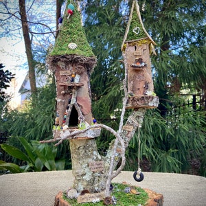 Peut inclure: Une maison de fée fantaisiste réalisée avec des matériaux naturels, comprenant deux tours avec des toits recouverts de mousse, une balançoire et le texte "All Fae's Welcome" et "Gorky Happy House".