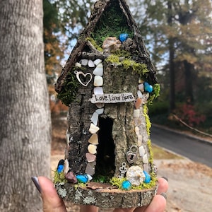 May include: A miniature fairy house made of bark and moss, decorated with small shells, stones, and glass beads. The house has a small door and a sign that reads "Love Lives Here".