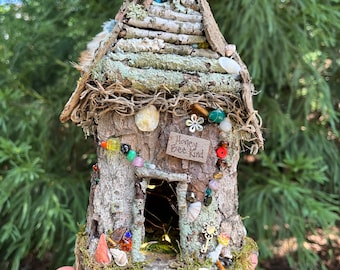 Könnte beinhalten: Ein Miniatur-Feenhaus aus Zweigen, Rinde und Moos, verziert mit bunten Perlen, Muscheln und einem Schild mit der Aufschrift "Happy Bee Kind".