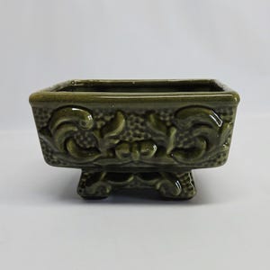 May include: A square olive green ceramic planter featuring a glossy finish and raised scrollwork relief patterns on all four sides. The vessel is supported by four short, ornate feet and has a textured background behind the embossed designs.