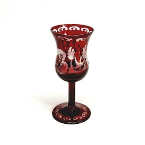 May include: A deep red glass goblet featuring intricate white etched floral and leaf patterns around the bowl and base. The stem is smooth and cylindrical, leading to a wide, circular foot. The rim is decorated with a repeating white scroll design.
