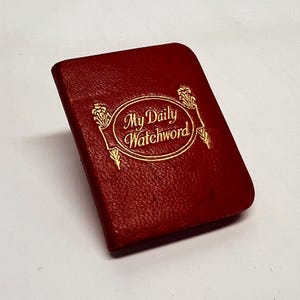 May include: A small, rectangular, red leather-bound book with the gold-colored text "My Daily Watchword" in an oval frame. The book has a textured surface and a slightly curved spine.